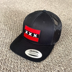 Puede incluir: Gorra de camionero negra con un parche de la bandera de Amsterdam rojo y blanco. La gorra tiene una parte trasera de malla negra y una visera negra. La gorra tiene una pegatina blanca en la parte delantera que dice "The Authentic Yupoong Snapback Yupoong Since 1974."