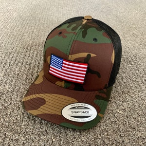 May include: A camouflage trucker hat with a black mesh back. The hat has a small American flag patch on the front and a white label with the text "The Authentic Yupoong Snapback Yupoong Since 1974".