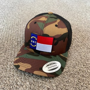 May include: A camouflage trucker hat with a black mesh back. The hat features a North Carolina state flag patch on the front. The hat has a black and silver sticker on the back that says "The Authentic Yupoong Snapback Since 1974."