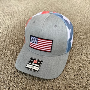 May include: A gray and blue baseball cap with a red, white, and blue American flag patch on the front. The cap has a mesh back and a white "Richardson" logo with the text "Est. 1970" and "Original 112 Trucker" on the side.