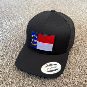 May include: A black and red North Carolina state flag patch on a black mesh trucker hat. The hat has a black brim and a black crown with a mesh back. The hat has a white label with the text "The Authentic Yupoong Snapback Yupoong Since 1974".