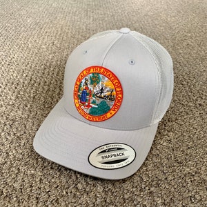 Può includere: Un cappello da camionista in rete grigio e bianco con una toppa che presenta il Grande Sigillo dello Stato della Florida. La toppa è rossa, gialla, verde e blu con il testo "Great Seal of the State of Florida" e "In God We Trust". Il cappello ha un'etichetta nera che dice "The Authentic Yupoong Snapback Yupoong Since 1974."
