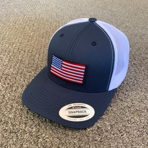 May include: A navy blue and white mesh baseball cap with an American flag patch on the front. The cap has a curved brim and a snap closure. The label on the back reads "The Authentic Yupoong Snapback Yupoong Since 1974."