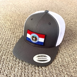 以下が含まれることがあります： 前面にミズーリ州の旗のパッチが付いたグレーとホワイトのトラッカーハット。帽子には「The Authentic Snapback」という黒いラベルに「Yupoong Since 1974」という文字が書かれています。