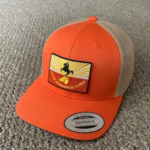 May include: Orange and tan trucker hat with a patch featuring the City of Jacksonville, Florida logo. The logo features a silhouette of a person riding a horse with a sunburst design. The hat has a mesh back and a snap closure. The hat also has a label that reads "The Authentic Yupoong Snapback Yupoong Since 1974."