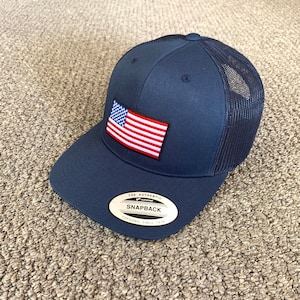 May include: A navy blue trucker hat with a red, white, and blue American flag patch on the front. The hat has a mesh back and a black sticker that says "The Authentic Snapback Yupoong Since 1974".
