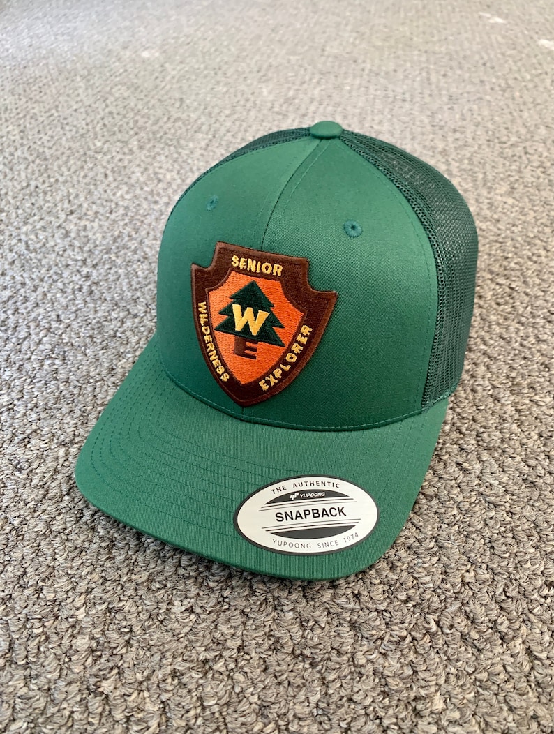 May include: A green and black mesh trucker hat with a brown leather patch that says "Senior Wilderness Explorer" and a white "W" inside. The hat also has a white label that says "The Authentic Yupoong Snapback Yupoong Since 1974".