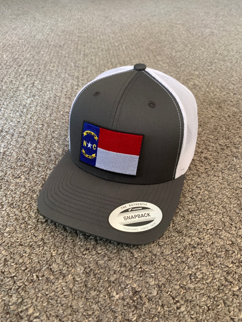 May include: A gray and white trucker hat with a North Carolina state flag patch on the front. The hat has a black "The Authentic Yupoong Snapback Yupoong Since 1974" label on the back.