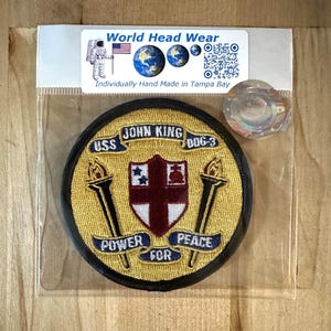 以下が含まれることがあります： 「USS JOHN KING DDG-3」と「POWER FOR PEACE」の文字が入った丸い刺繍パッチ。パッチの中央には赤い十字と2つのトーチが金色の背景に描かれています。パッチは透明なビニール袋に梱包されています。