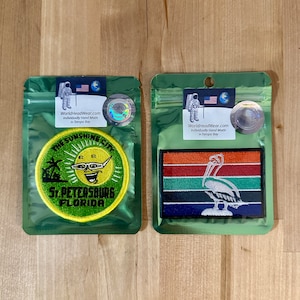 May include: Two embroidered patches in clear plastic bags. The first patch is round and features a smiling sun with sunglasses and the text "The Sunshine City St. Petersburg Florida". The second patch is rectangular and features a white pelican on a multi-colored striped background.