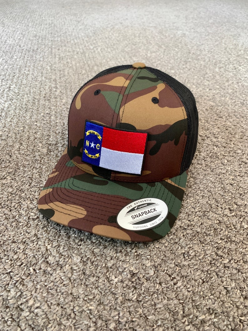 May include: A camouflage trucker hat with a black mesh back. The hat features a North Carolina state flag patch on the front. The hat has a black and silver sticker on the back that says "The Authentic Yupoong Snapback Since 1974."