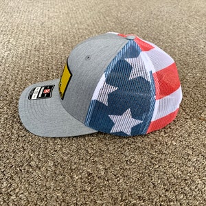 May include: A gray and white baseball cap with a mesh back. The front of the cap has a yellow patch with black text and the back has a red, white, and blue American flag design.