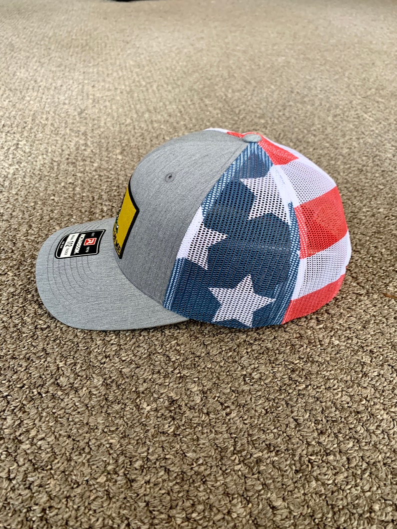 May include: A gray and white baseball cap with a mesh back. The front of the cap has a yellow patch with black text and the back has a red, white, and blue American flag design.