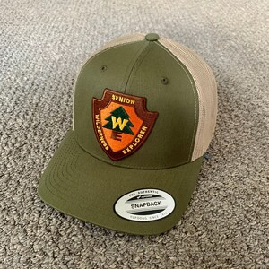 May include: A green and tan trucker hat with a patch that says "Senior Wilderness Explorer" and a white label that says "The Authentic Snapback Yupoong Since 1974".