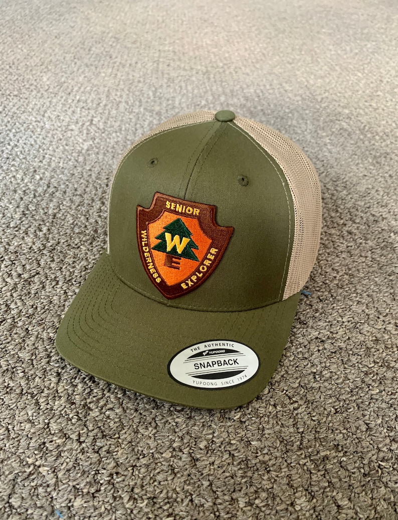 May include: A green and tan trucker hat with a patch that says "Senior Wilderness Explorer" and a white label that says "The Authentic Snapback Yupoong Since 1974".