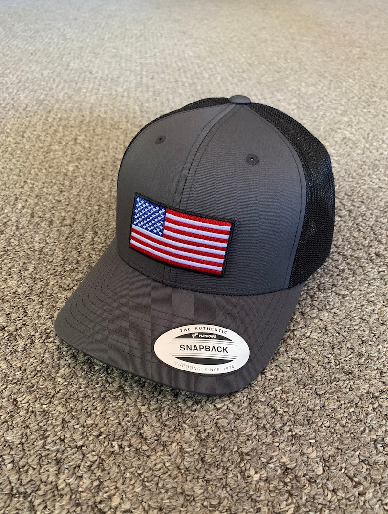 May include: A gray and black mesh baseball cap with a small American flag patch on the front. The cap has a black mesh back and a gray front with a black brim. The cap has a white label on the front that says "The Authentic Yupoong Snapback Yupoong Since 1974."