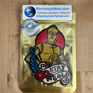 May include: A gold embroidered patch featuring a robot character with a red background and the text "Stay Gold". The patch is surrounded by a blue and green floral design.