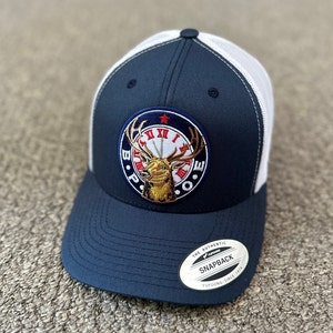 May include: A navy blue and white trucker hat with a white mesh back. The hat features a large embroidered patch with a red star, a clock face, and a deer with antlers. The text "B.U.N.I.E" is embroidered around the clock face. The hat also has a white label with the text "The Authentic Yupoong Snapback Since 1974".