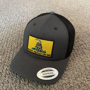 May include: A gray and black mesh trucker hat with a yellow patch featuring a snake and the text "Don't Tread On Me". The hat has a black mesh back and a gray front with a black brim. The hat has a white sticker on the front that says "The Authentic Yupoong Snapback Yupoong Since 1974".