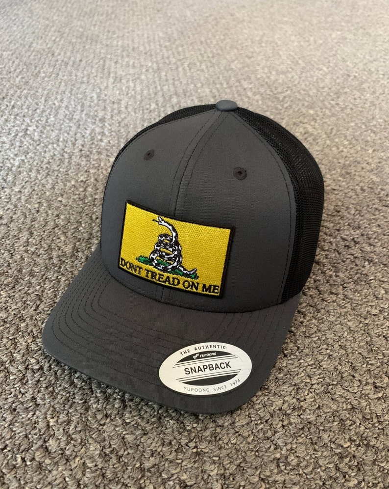 May include: A gray and black mesh trucker hat with a yellow patch featuring a snake and the text "Don't Tread On Me". The hat has a black mesh back and a gray front with a black brim. The hat has a white sticker on the front that says "The Authentic Yupoong Snapback Yupoong Since 1974".