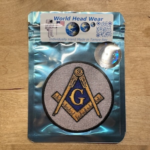 Op de afbeelding: Een geborduurde grijze en blauwe patch met een kompas en een geodriehoek, een letter G in het midden, en de tekst "World Head Wear" en "Individually Hand Made in Tampa Bay" op een wit label.