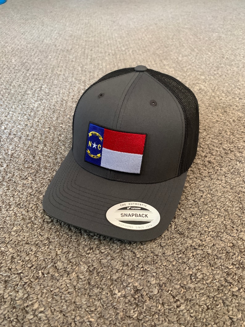 May include: A gray and black mesh baseball cap with a North Carolina state flag patch on the front. The cap has a black mesh back and a gray front with a black brim. The cap has a sticker on the back that says "The Authentic Yupoong Snapback Yupoong Since 1974".