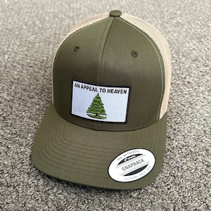 May include: A green and beige trucker hat with a patch that says "An Appeal to Heaven" with a small green pine tree graphic. The hat has a white label that says "The Authentic Yupoong Snapback Yupoong Since 1974".