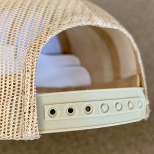 May include: A beige mesh trucker hat with a white adjustable strap and six holes for size adjustment. The strap has a small logo.