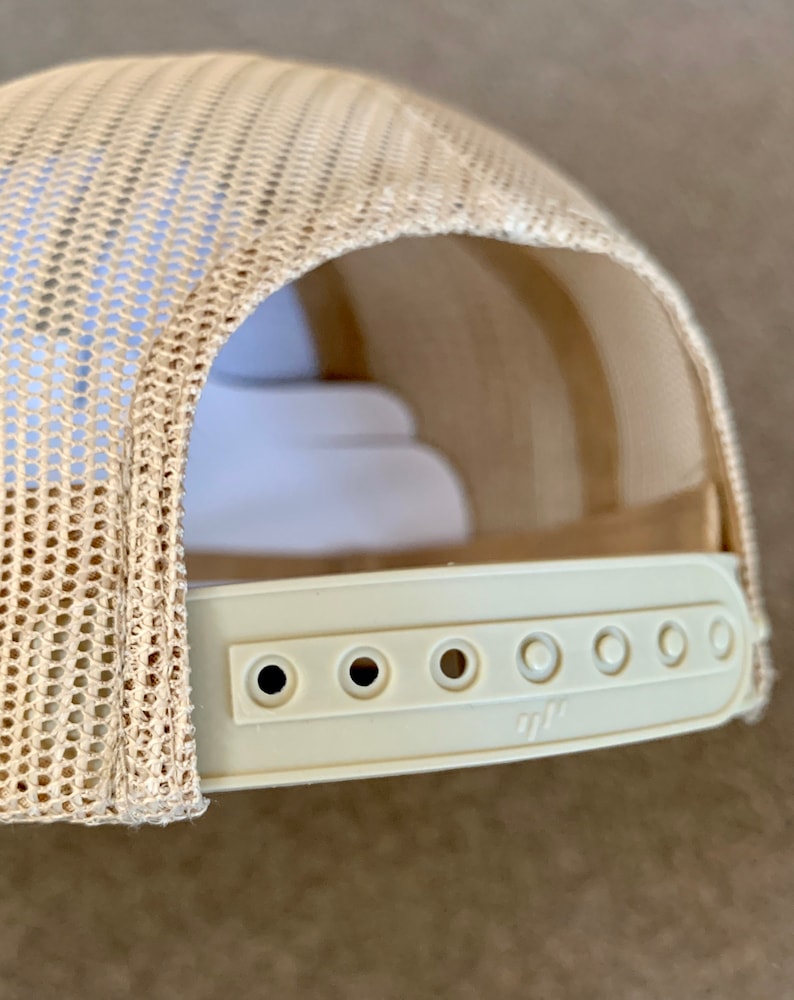 May include: A beige mesh trucker hat with a white adjustable strap and six holes for size adjustment. The strap has a small logo.