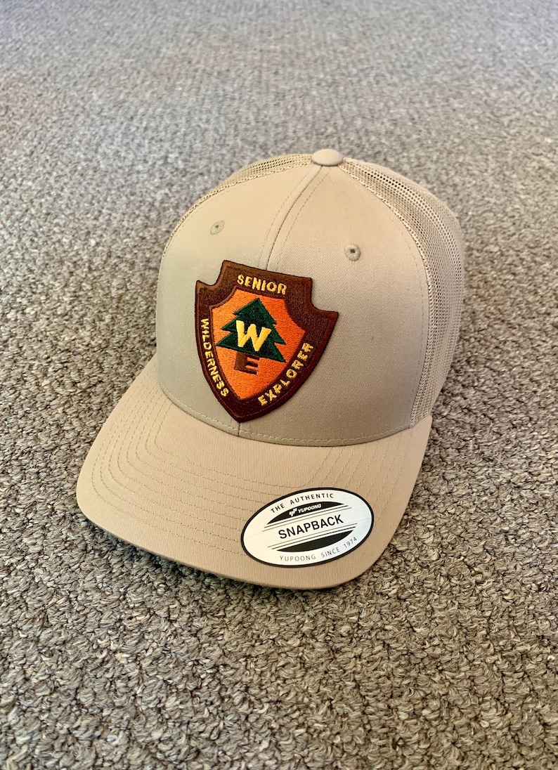 May include: A tan and brown mesh baseball cap with a patch that says "Senior Wilderness Explorer" and a logo that says "The Authentic Yupoong Snapback Yupoong Since 1974".