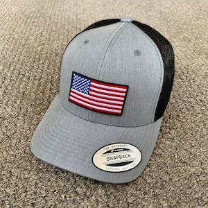 May include: A gray and black baseball cap with a mesh back. The cap has an American flag patch on the front and a "The Authentic Yupoong Snapback Yupoong Since 1974" label on the side.