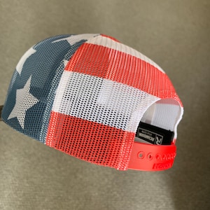 May include: Red, white, and blue mesh trucker hat with a star on the front. The hat has a red adjustable strap with the word "Richardson" printed on it.
