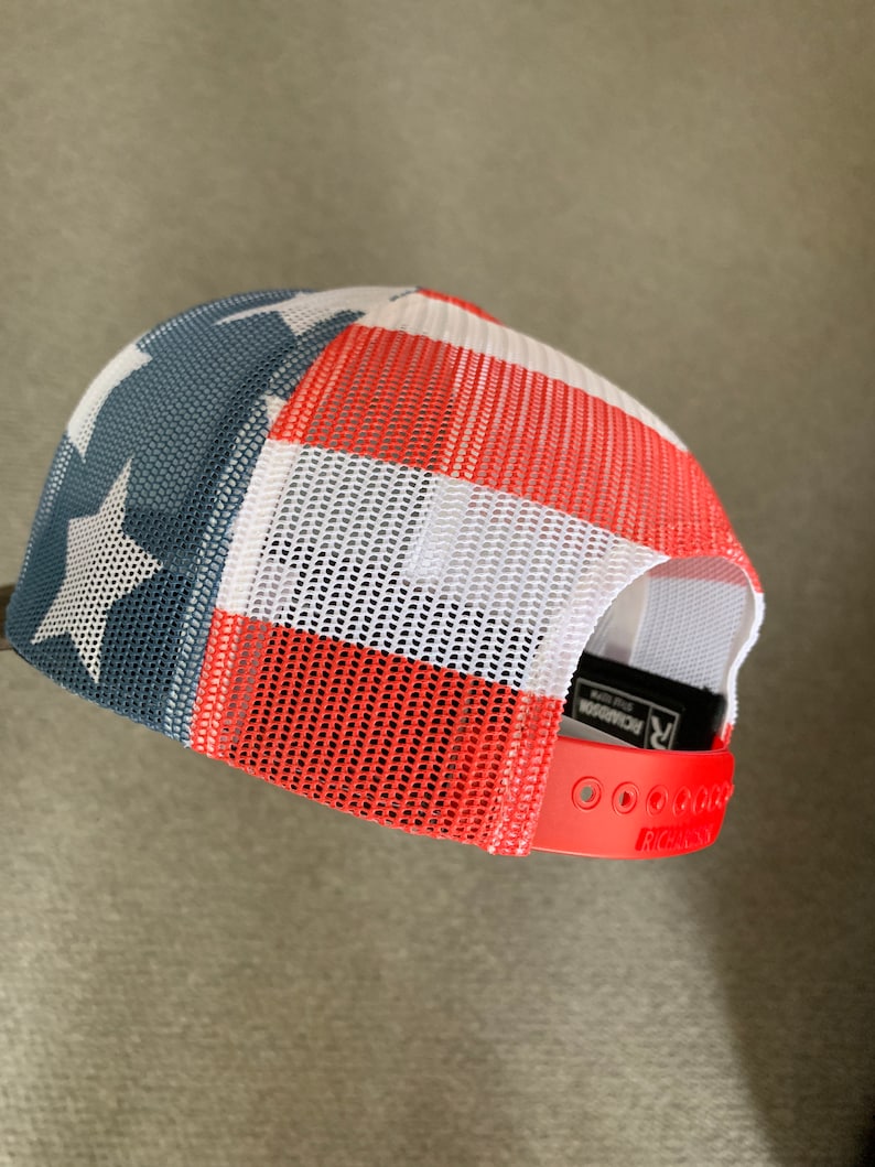May include: Red, white, and blue mesh trucker hat with a star on the front. The hat has a red adjustable strap with the word "Richardson" printed on it.