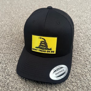 May include: Black trucker hat with a yellow patch featuring a coiled snake and the text "Don't Tread On Me". The hat has a black mesh back and a silver sticker that reads "The Authentic Yupoong Snapback Yupoong Since 1974".