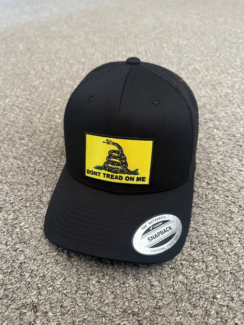 May include: Black trucker hat with a yellow patch featuring a coiled snake and the text "Don't Tread On Me". The hat has a black mesh back and a silver sticker that reads "The Authentic Yupoong Snapback Yupoong Since 1974".
