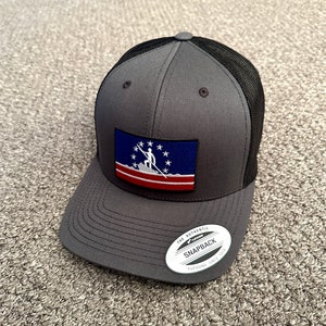 May include: A gray and black trucker hat with a blue, white, and red patch featuring a person holding a spear. The patch has a white star pattern and the text "THE AUTHENTIC YUPOONG SNAPBACK SINCE 1974".