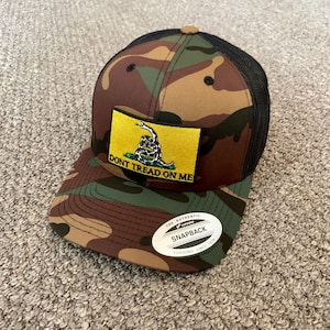 May include: A camouflage trucker hat with a black mesh back. The hat features a yellow patch with a coiled snake and the text "Don't Tread On Me". The hat also has a black and white sticker that says "The Authentic Yupoong Snapback Yupoong Since 1974".