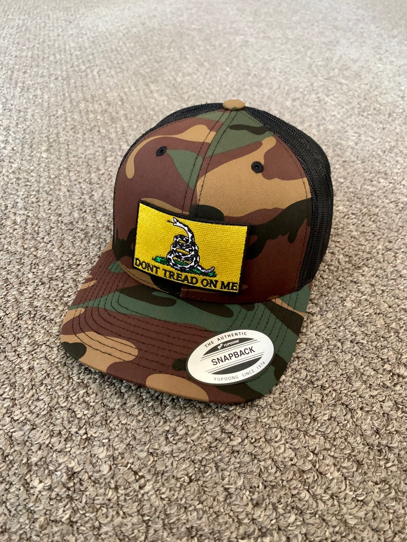 May include: A camouflage trucker hat with a black mesh back. The hat features a yellow patch with a coiled snake and the text "Don't Tread On Me". The hat also has a black and white sticker that says "The Authentic Yupoong Snapback Yupoong Since 1974".