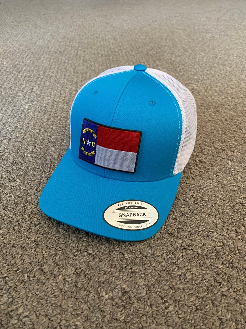 May include: A turquoise blue and white trucker hat with a North Carolina state flag patch. The hat has a white mesh back and a black "The Authentic Snapback Yupoong Since 1974" label.