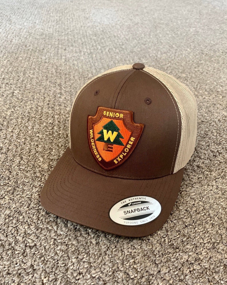 May include: Brown and beige trucker hat with a patch that says "Senior Wilderness Explorer" with a stylized tree and the letter "W". The hat has a brown brim and a beige mesh back. The hat has a label that says "The Authentic Snapback Yupoong Since 1974".