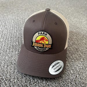Puede incluir: Gorra de camionero marr&oacute;n y beige con un parche de logotipo de Jurassic Park Ranger. El parche presenta un c&iacute;rculo amarillo con el texto "Park" en la parte superior, "Jurassic Park" en el medio y "Ranger" en la parte inferior. Un dinosaurio rojo est&aacute; dentro del c&iacute;rculo. La gorra tiene una parte delantera marr&oacute;n y una parte trasera de malla beige. La gorra tiene una pegatina blanca en el lateral que dice "The Authentic Snapback Yupoong Since 1974."