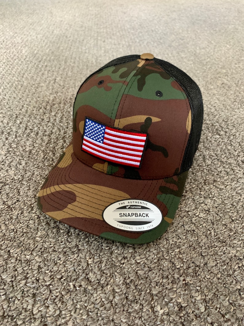 May include: A camouflage trucker hat with a black mesh back. The hat has a small American flag patch on the front and a white label with the text "The Authentic Yupoong Snapback Yupoong Since 1974".