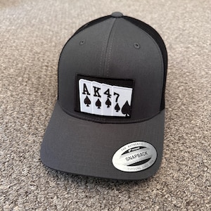 Może przedstawiać: Ciemnoszara czapka typu snapback z czarną siateczką z tyłu i prostokątną naszywką z przodu. Naszywka przedstawia cztery białe karty do gry z literami A, K, 4, 7 oraz czarnymi symbolami pik. Na daszku znajduje się srebrna naklejka.