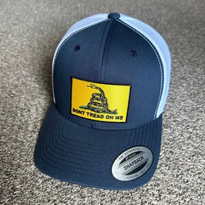 May include: A navy blue and white trucker hat with a yellow patch featuring a coiled snake and the text "Don't Tread On Me". The hat has a mesh back and a YUPOONG snapback closure.