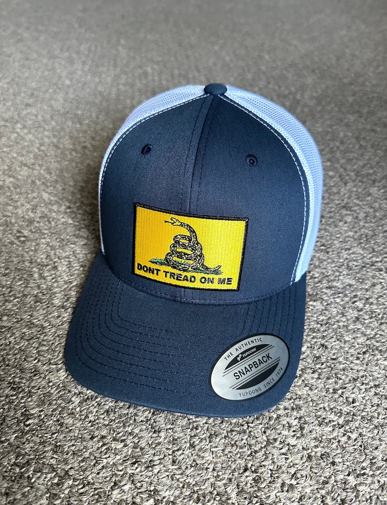 May include: A navy blue and white trucker hat with a yellow patch featuring a coiled snake and the text "Don't Tread On Me". The hat has a mesh back and a YUPOONG snapback closure.