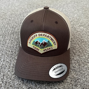 May include: A brown and tan baseball cap with a curved brim. The cap features a patch with the words "SHERIFF DEPARTMENT TWIN PEAKS" and an image of mountains and trees. The cap has a snapback closure.