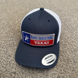 Puede incluir: Una gorra de camionero azul marino y blanca con un parche rojo, blanco y azul que dice "Don't mess with Texas". La gorra tiene una malla blanca en la parte posterior y una correa ajustable negra con una etiqueta "The Authentic Yupoong Snapback".