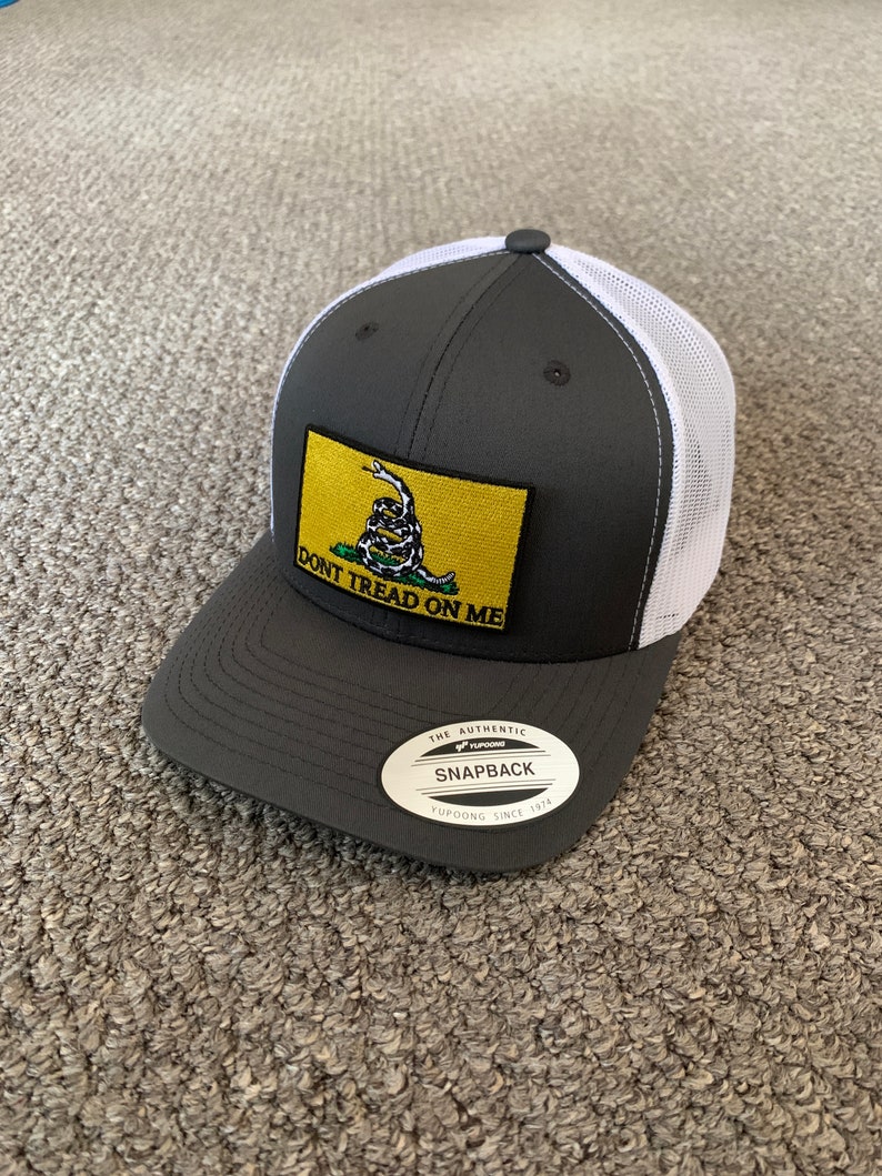 May include: A gray and white trucker hat with a yellow patch that says "Don't Tread On Me" and a snake with a yellow background. The hat has a black label that says "The Authentic Yupoong Snapback Yupoong Since 1974".