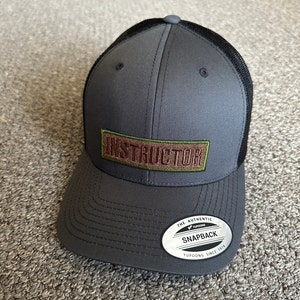 May include: Gray and black mesh trucker hat with a brown patch that says "INSTRUCTOR" in white letters. The hat has a black mesh back and a gray front with a black brim. The hat has a white label that says "THE AUTHENTIC YUPOONG SNAPBACK YUPOONG SINCE 1974".
