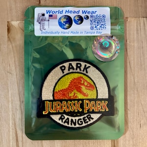 Könnte beinhalten: Ein weißer, runder Patch mit einem gelben Kreis in der Mitte. Der Kreis zeigt die Silhouette eines roten Dinosauriers. Der Text "PARK" befindet sich über dem Kreis, "JURASSIC PARK" unter dem Kreis und "RANGER" in einem schwarzen Kreis unter dem Text.